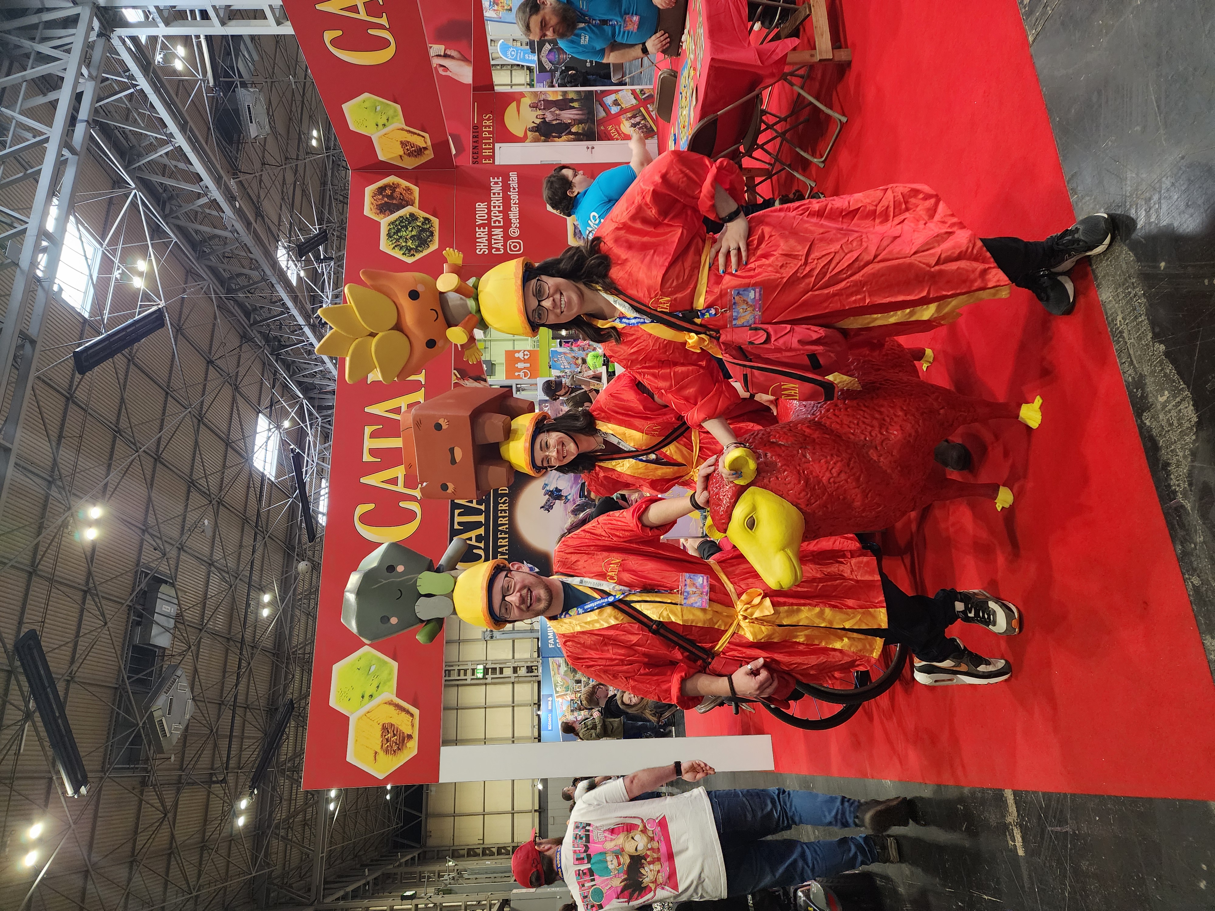A group of three people standing in red robes with the CATAN logo, with yellow helmets on their heads, with a different CATAN resource on top of the helmet. 