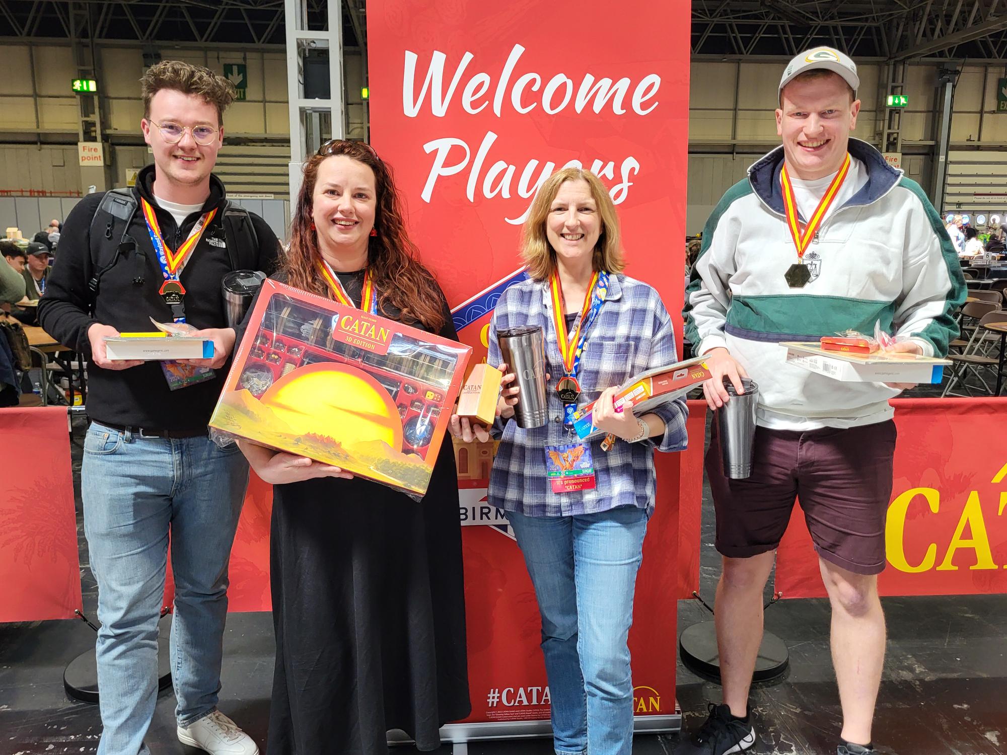 The four finalists of the CATAN UK National Championship stand together in an expo hall, standing in front of a CATAN banner. 