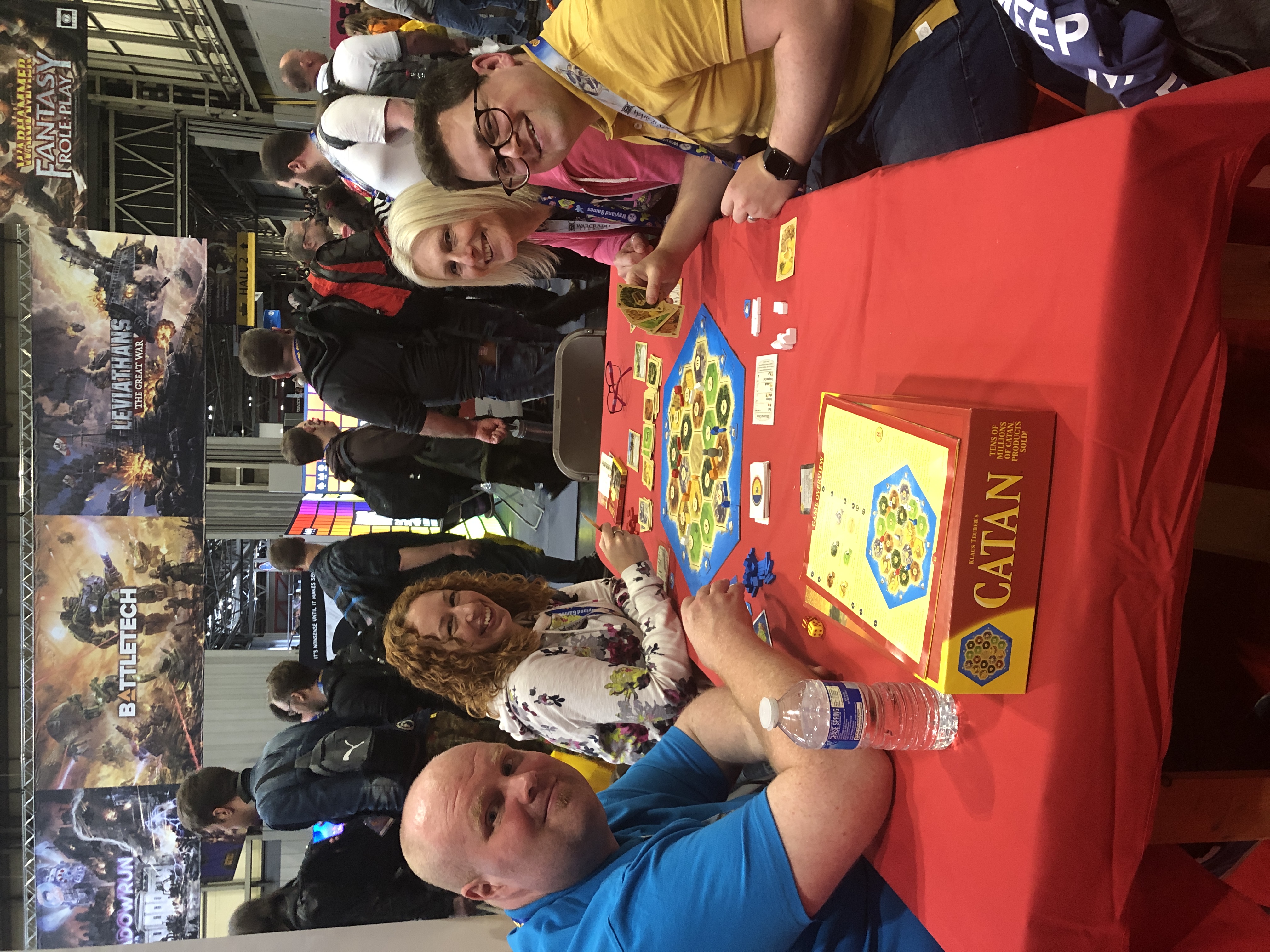 A group of four people smiling for the camera while sitting around a table playing CATAN.