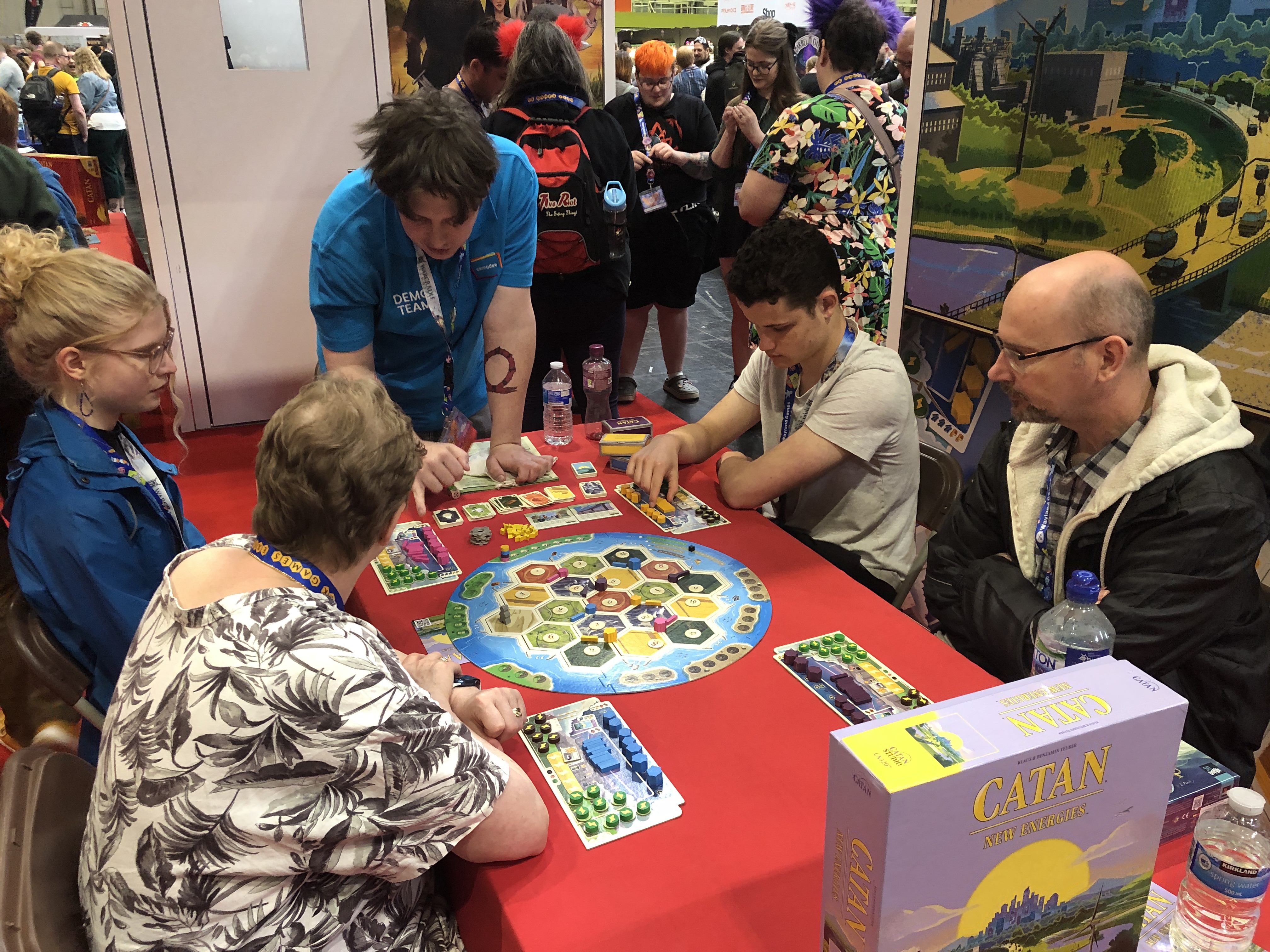 A group of 4 people sitting at a table as a blue shirted Demo teacher helps them learn CATAN – New Energies.