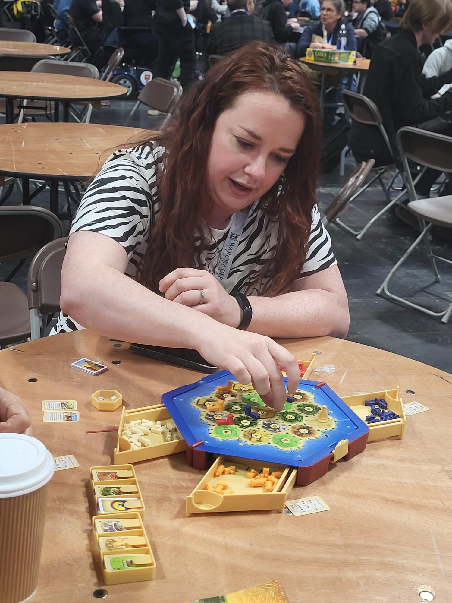 A woman with red hair is moving a piece on a small CATAN – Traveler board.