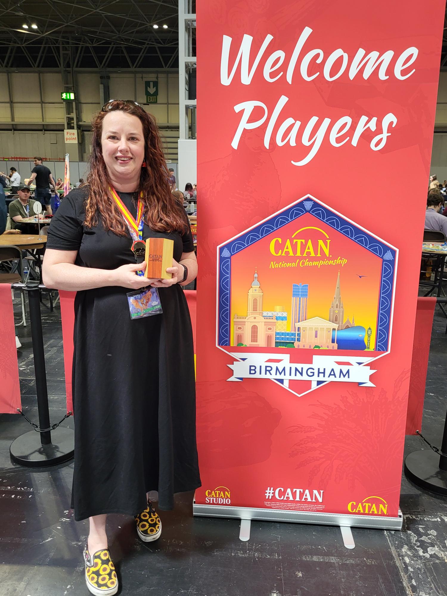 Jess Burns is pictured wearing a black dress and holding a small wooden trophy. She's standing in a expo hall next to a red banner that says CATAN Championship Birmingham. 