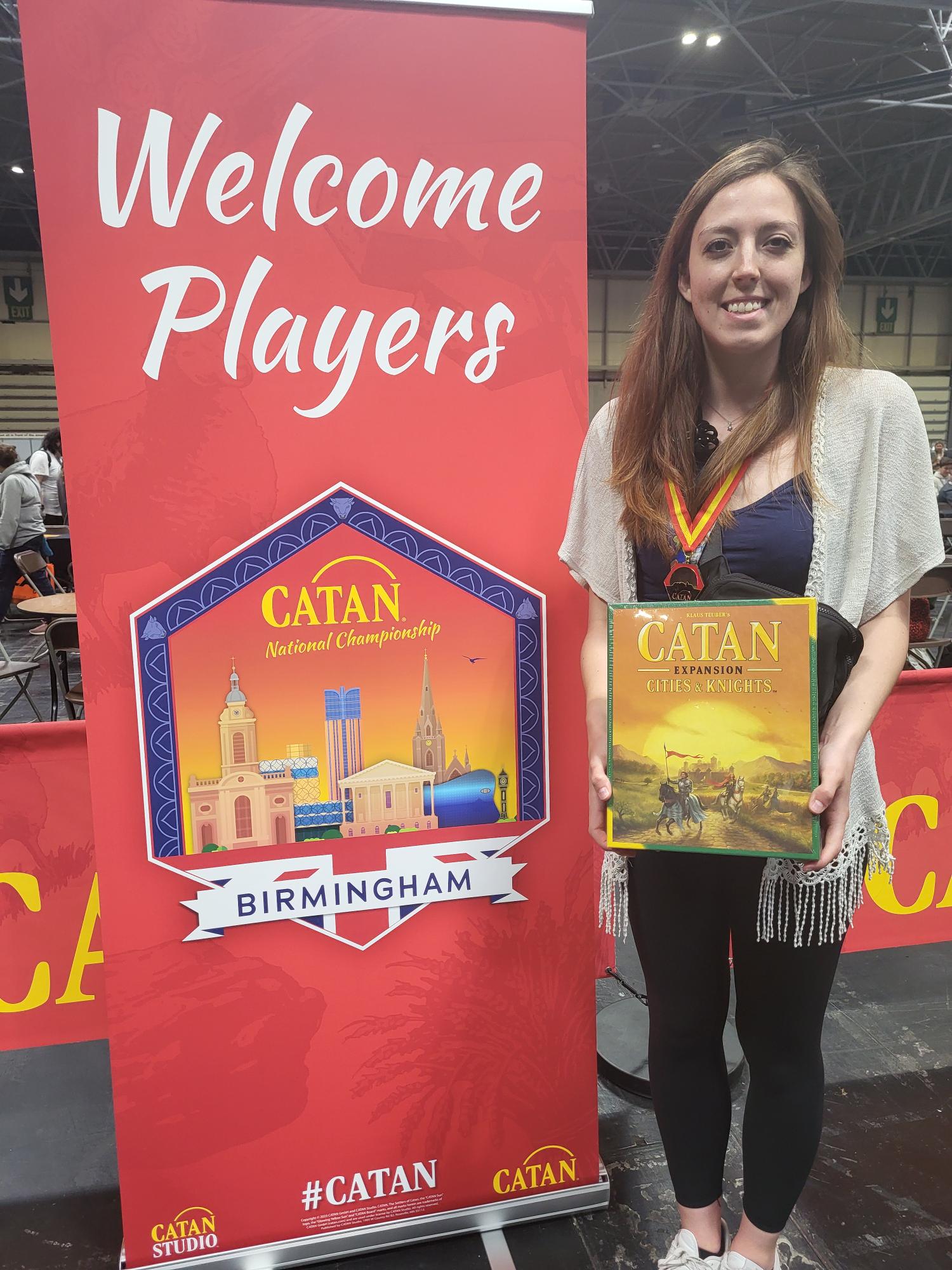 A picture of Katrina Tripple holding a CATAN – Cities & Knights box while standing on the right to a red banner that says welcome players, with text below saying CATAN Championships Birmingham. 