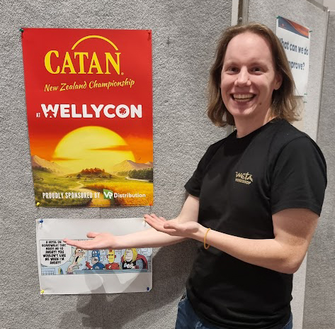 A man with longer blonde hair stands and smiles while gesturing to a red poster that says CATAN New Zealand Championship, Wellycon. 