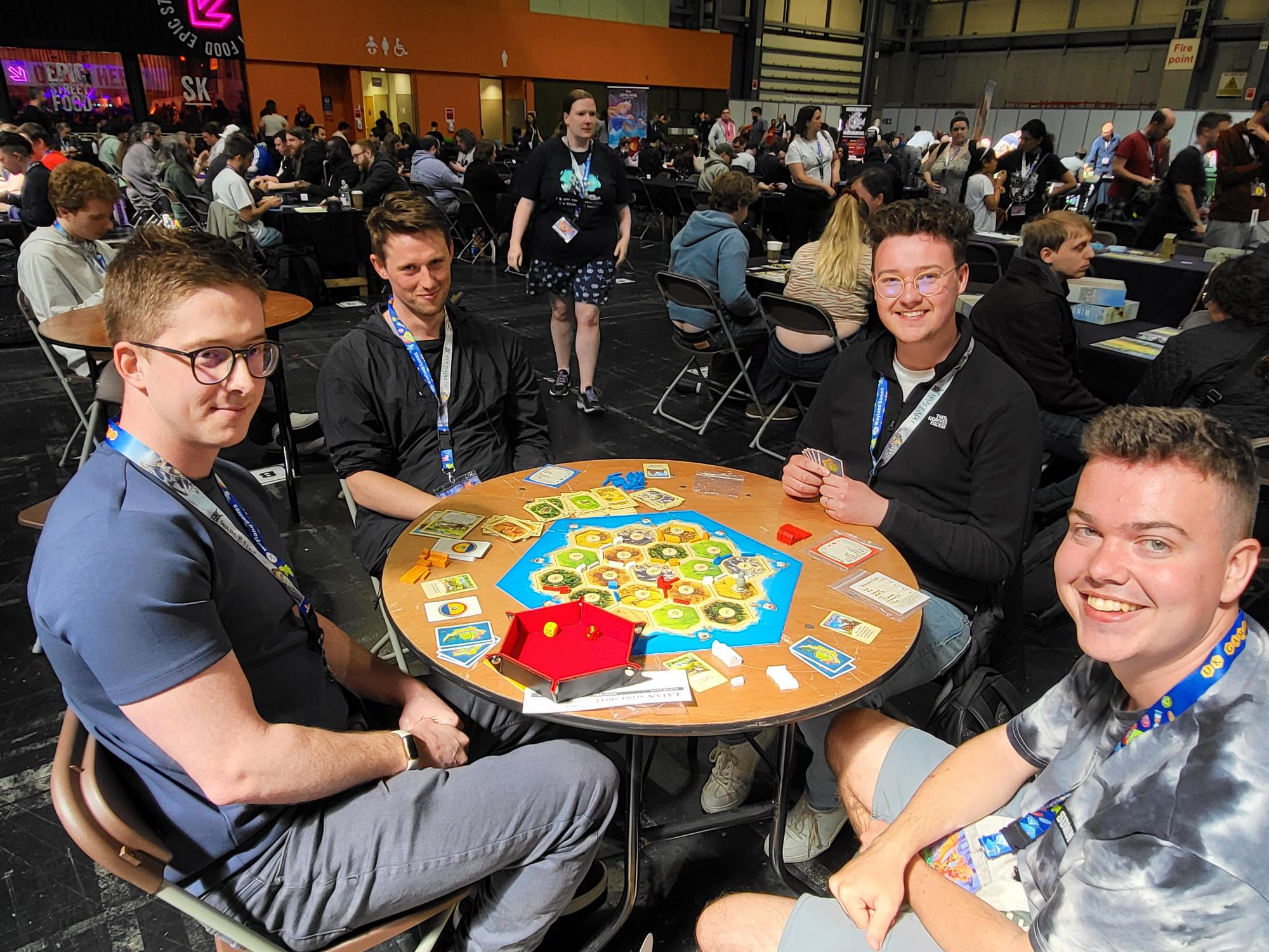 A group of people sit around a round table playing CATAN while smiling for the camera. 